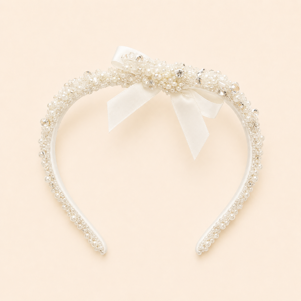 A white headband embellished with delicate pearls, clear rhinestones, and a central white satin bow, set against a solid pale peach background.