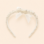 A white headband embellished with delicate pearls, clear rhinestones, and a central white satin bow, set against a solid pale peach background.