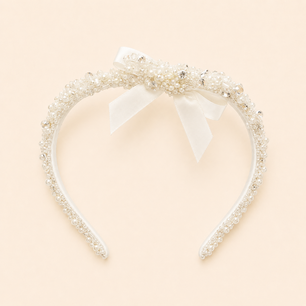 A white headband embellished with delicate pearls, clear rhinestones, and a central white satin bow, set against a solid pale peach background.